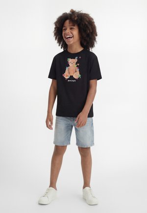 Niño sonriente con cabello rizado que lleva una camiseta negra con estampado de oso de peluche, pantalones cortos de mezclilla claros y zapatillas blancas, de pie sobre un fondo blanco.