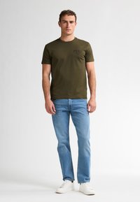 Olivgrünes Baumwoll-T-Shirt mit kurzen Ärmeln, auf der Brust mit einem dunkelblauen Logo bedruckt, kombiniert mit hellblauen Jeans und weißen Sneakers.