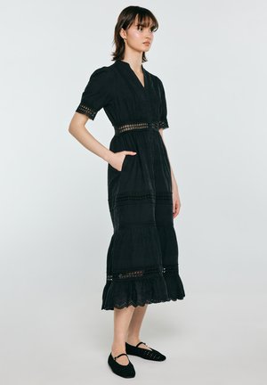 Kobieta stojąca w czarnej sukience midi z koronkowymi detalami, krótkimi rękawami i kieszeniami, w czarnych płaskich butach, na jednolitym jasnym tle.