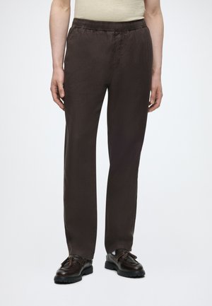 Personne portant un pantalon droit marron foncé avec une taille élastique et des chaussures en cuir noires à lacets sur un fond uni.