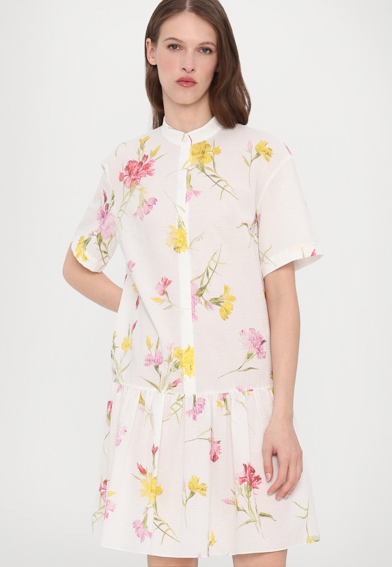 Robe blanche à manches courtes avec un motif floral en rose et jaune, présentant une fermeture à boutons à l'avant et un ourlet à volants.