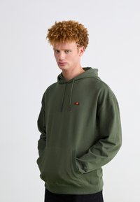 Sudadera verde de tejido suave, con un bolsillo frontal y cordones. Pequeño logotipo de color en el pecho. Ajuste relajado con dobladillo acanalado.