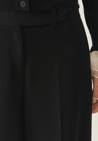 Zwarte broeken met een getailleerde pasvorm, voorzien van een vlakke tailleband, zijzakken en een knoopdetail. Gladde textuur met een gepolijste afwerking.