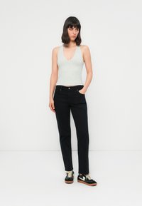 Kvinne som står med den ene hånden i lommen, iført en lysegrå ermeløs topp, sorte jeans og svarte joggesko med hvite snørebånd.