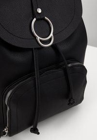 Czarny plecak teksturowany z zaokrągloną klapą, metalowymi pierścieniami i zaciąganym zamknięciem; posiada przednią kieszeń na zamek.