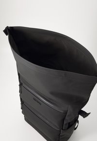 Backpack with a tall, rectangular shape, made of matte black material. Features two front zippered pockets and adjustable side straps.