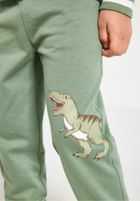 Zielone dresowe spodnie z bawełny z nadrukowanym dinozaurem na lewej udzie. Materiał jest miękki, ma gładką teksturę i elastyczny pas.