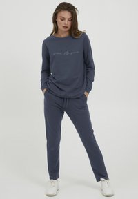 Felpa blu navy con il testo "Eleganza Informale", abbinata a pantaloni jogger coordinati. Entrambi realizzati in un tessuto morbido e testurizzato. Sneakers bianche.