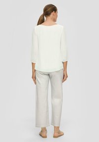 Blouse blanche côtelée à manches trois-quarts, associée à un pantalon large gris clair. Cheveux blonds en queue de cheval, sandales visibles.