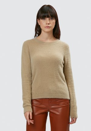 Beige, gestructureerde gebreide trui met lange mouwen en een ronde halslijn, gedragen met bruine leren broek, tegen een neutrale achtergrond.