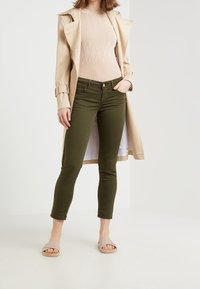 Olijfgroene skinny jeans met een knoopsluiting en voorzakken, gecombineerd met een beige geribde trui en crèmekleurige open-teensandalen.