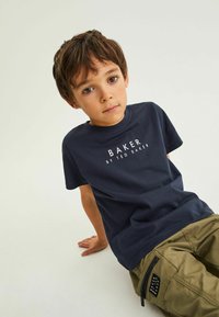 T-paita tummansinisestä puuvillasta, jossa on valkoinen "BAKER BY TED BAKER" -teksti. Lyhyet hihat, pyöreä pääntie; yhdistetty khaki-värisiin cargo-housuihin, joissa on sivutasku.