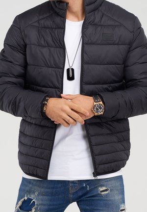 Man draagt een zwart doorgestikt jack over een wit shirt, versleten blauwe spijkerbroek, polshorloge, armband en een zwarte dogtagketting.