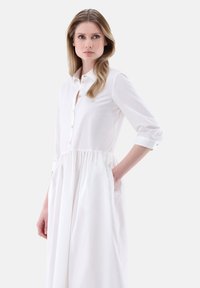 Abito bianco a camicia con corpino aderente, vita arricciata, maniche lunghe e tasche laterali nascoste. Tessuto liscio e leggero.