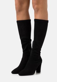 Bottes noires en daim montant jusqu'au genou avec un bout pointu, un talon épais, une texture lisse et un détail de fermeture éclair latérale.