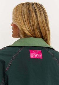 Zelená bunda s velkým límcem, růžovým prošitím a výraznou růžovou nášivkou s potiskem vzadu. Texturovaná tkanina.