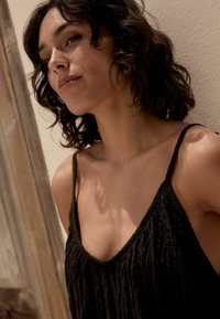 Haut noir texturé avec des fines bretelles, à la coupe ample et à encolure dégagée, sur fond de mur clair.