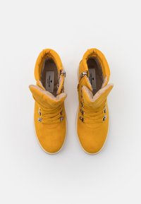 Bottes cheville en daim jaune avec doublure en fourrure, semelles plates en caoutchouc, lacets réglables et détails en métal sur le côté pour un enfilage facile.