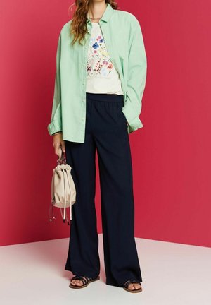 Lichtgroen overhemd over een wit T-shirt met bloemenprint, gecombineerd met donkergroene wijde broek en zwarte sandalen met banden. Beige tas met trekkoord vastgehouden.