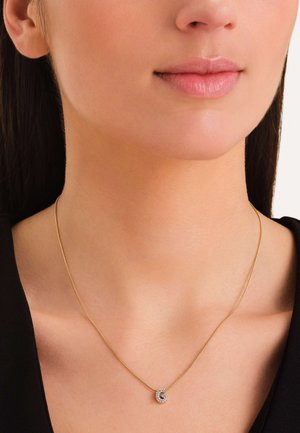 Collar de oro con un colgante en espiral que presenta piedras preciosas transparentes, reposando sobre un cuello desnudo, contra una blusa negra. Diseño simple y elegante.