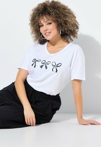 T-shirt blanc à manches courtes avec un col en V, arborant trois graphismes de nœuds noirs à l'avant. Assorti à une jupe noire à taille élastique.