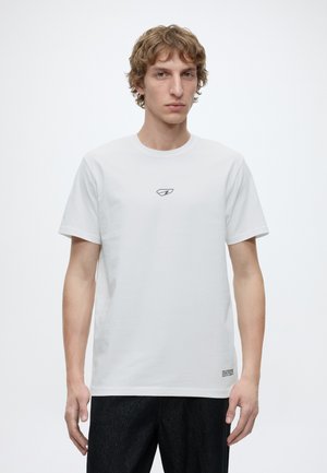 Jeune homme aux cheveux bouclés portant un t-shirt blanc uni avec un petit logo sur la poitrine et un pantalon foncé sur fond blanc.