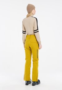Beige Langarmhemd mit schwarzen Streifen an den Ärmeln, kombiniert mit gelben Skihosen mit Reißverschlusstaschen und seitlichen Schlitzen. Strickmütze in Beige.