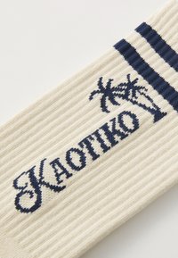 Strikkede creme sokker med ribbet tekstur, marineblå striber og broderet "KAOTIKO" med palme grafik.