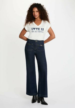 Donna con capelli ricci che indossa una maglietta bianca con la scritta "L'amore è bello", jeans scuri a gamba larga e vita alta, e stivaletti neri con tacco.