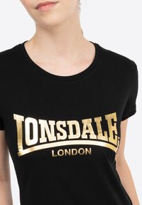 Melnā kokvilnas T-kreklu ar lielu zelta folijas apdruku "LONSDALE LONDON" uz krūšu, klasiskā apaļā kakla un īsajām piedurknēm.