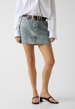 Femme portant une mini-jupe en jean bleu délavé avec une ceinture marron foncé, une chemise blanche à manches longues et des sandales à talons marron.