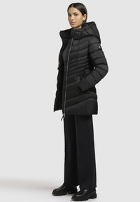 Sort pufferjakke med lynlås, hætte og quiltet design; har sidelommer og en glat tekstur. Sammen med sorte bukser og støvler.