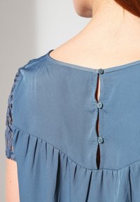Blå bluse med knaplukning bagpå, der har et plisseret design og blondeærmer. Glat stof med en let tekstur.