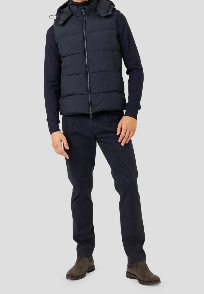 Chaleco acolchado azul marino con cremallera frontal y capucha desmontable, llevado sobre una camisa azul marino de manga larga, combinado con pantalones oscuros y botas marrones.