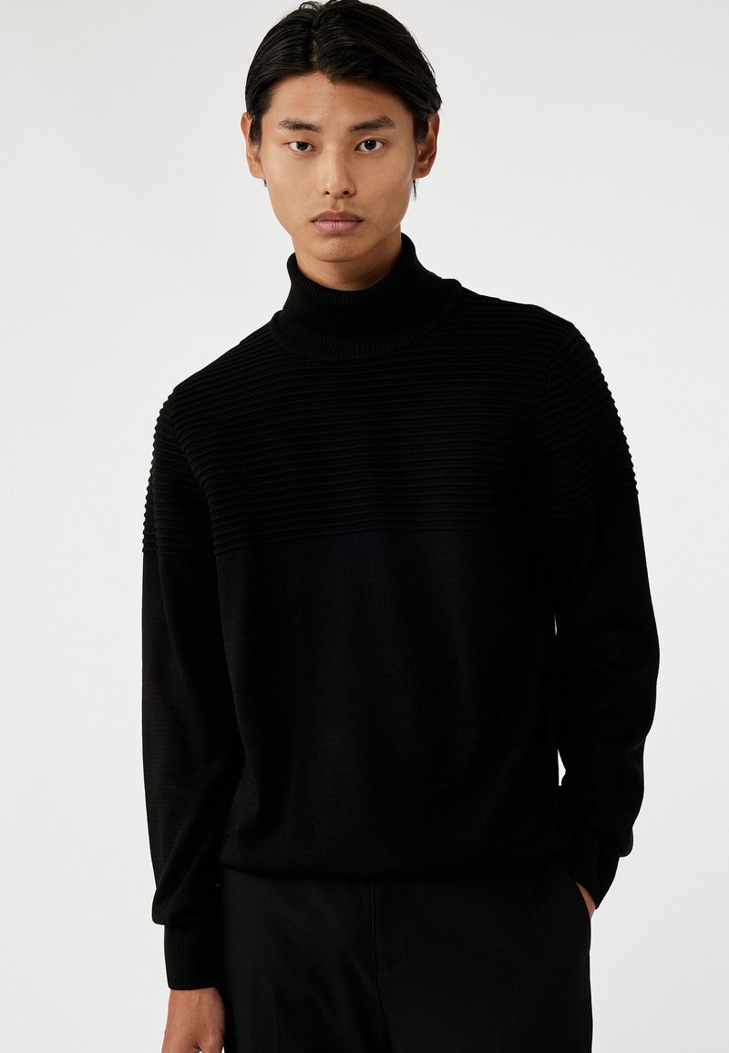 Suéter negro de cuello alto con textura acanalada en la parte superior y cuerpo inferior liso. Mangas largas, corte holgado y diseño minimalista.