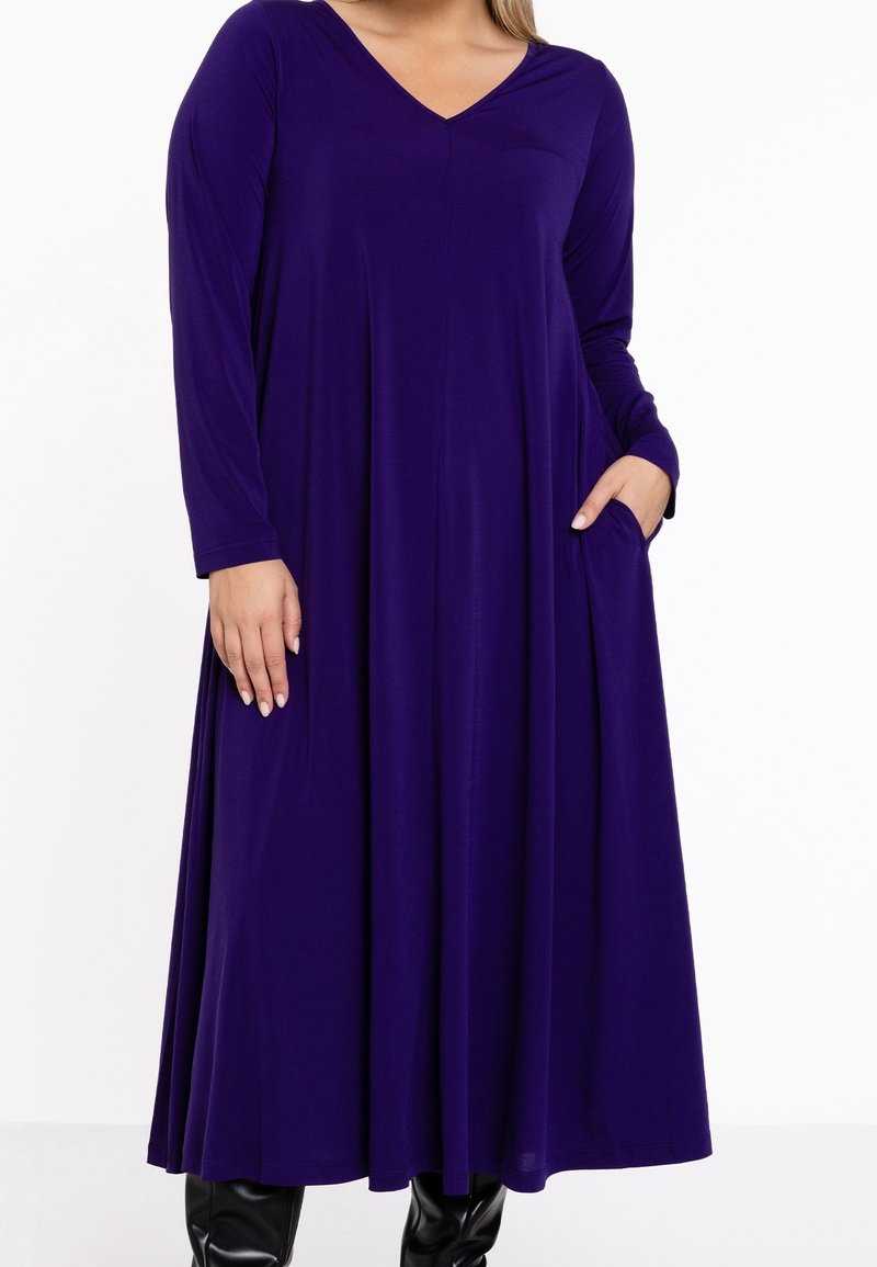 Robe à manches longues violet foncé avec un col en V et une jupe évasée, dotée de poches latérales, portée avec des bottes noires.