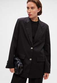 Blazer negru cu sclipiri subtile, rever tăiat, două buzunare frontale și nasturi argintii. Asortat cu o geantă pufosă neagră.