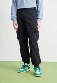 Mustat nailoniset cargo-housut joustavalla vyötärönauhalla, lyhyet lahkeet, sivutaskut ja hillitty Nike-logo, yhdistettynä vihreisiin ja valkoisiin lenkkareihin.