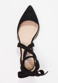 Scarpa ballerina nera con punta affilata, cinturini incrociati alla caviglia e dettagli a fiocco su una soletta di un beige chiaro, su sfondo bianco.