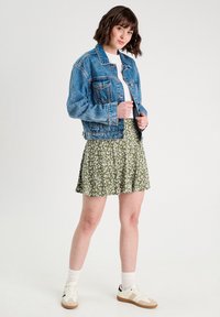 Veste en denim avec une coupe décontractée, associée à une mini-jupe florale verte et un t-shirt blanc, complétée par des chaussures de sport blanches et des chaussettes.