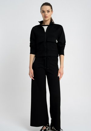 Kobieta w czarnej kurtce na zamek z dużymi przednimi kieszeniami, szerokich czarnych spodniach i czarno-białych trampkach, stojąca na tle jasnego, jednolitego tła.