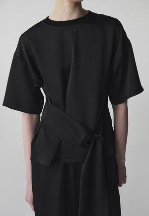 Persoon draagt een losse zwarte top met korte mouwen en een brede riem die in een lus om de taille is gebonden, tegen een effen grijze achtergrond.