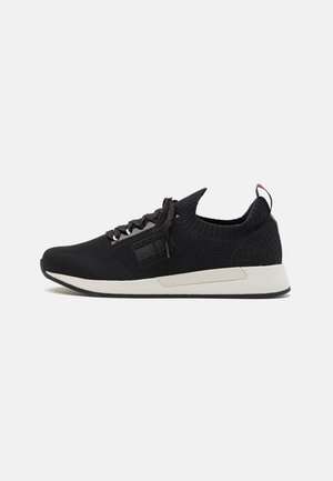 Czarny dziany niski sneaker z białą teksturowaną podeszwą, czarnymi sznurówkami i pętelką na pięcie, pokazany z profilu na jednolitym tle.