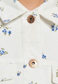 Hvit skjorte med små blå blomstertrykk, med krage og en kobberknapp med gravert tekst. Myk teksturert stoff.