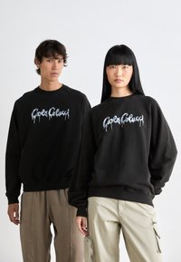 Zwarte crewneck sweatshirts hebben een druipend, gestileerd logo aan de voorkant. Modellen dragen beige en lichtgroene onderkleding. Gladde stof textuur.