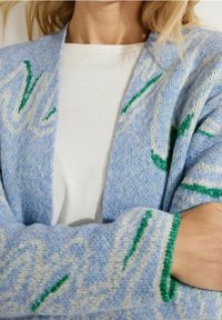 Helle blaue Strickjacke mit grünen und weißen abstrakten Mustern, weicher Textur, offene Vorderseite und überschnittenen Schultern, über einem weißen Hemd getragen.