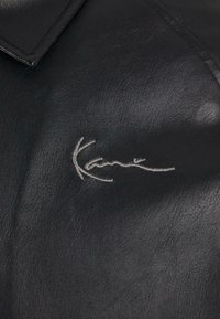 Sort læderstof med en glat tekstur og en krave. Har en sølv broderet signatur.