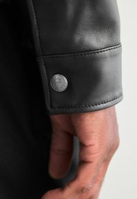 Schwarzlederjacke mit einem Bündchen, das einen Metallschnappverschluss mit eingraviertem Branding, gestickte Details und eine glatte Textur aufweist.