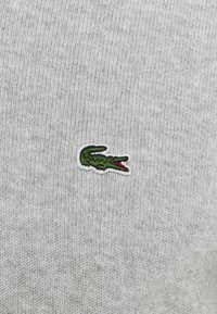 Jasnoszary sweter dziany z małym, haftowanym zielonym logo krokodyla z czerwonymi akcentami, umieszczonym na lewej stronie klatki piersiowej.