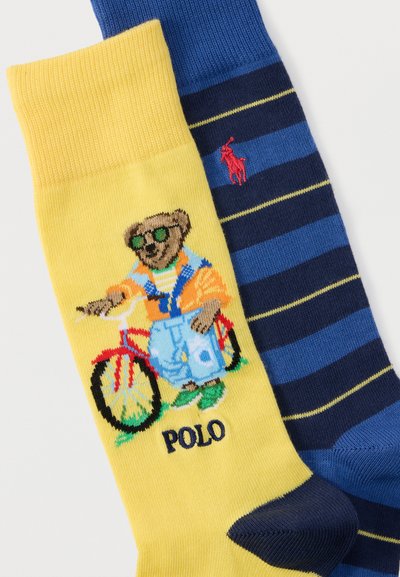 Κίτρινη κάλτσα με μια αρκούδα που φορά γυαλιά ηλίου και οδηγεί ένα κόκκινο ποδήλατο, συνδυασμένη με μια μπλε και ναυτικό ριγέ κάλτσα με κόκκινο λογότυπο αλόγου.